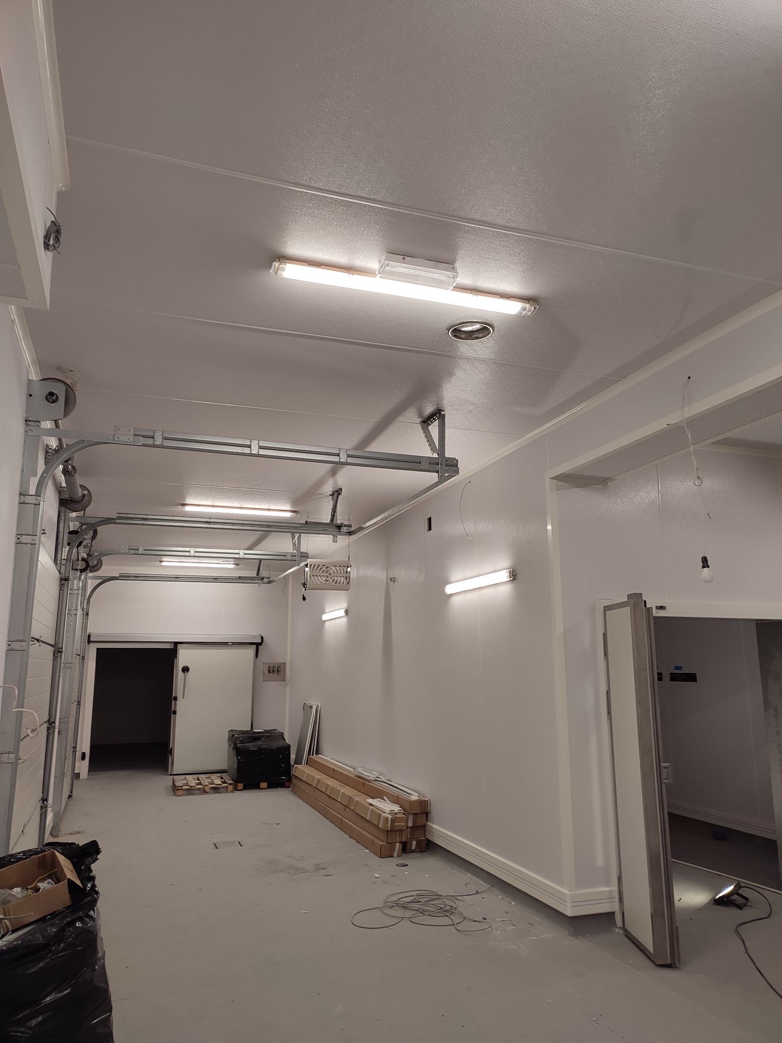 Wnętrze magazynu chłodniczego z zamontowanym oświetleniem liniowym, widoczne szyny jezdne bramy przesuwnej, przewody elektryczne na podłodze i elementy konstrukcyjne sufitu.