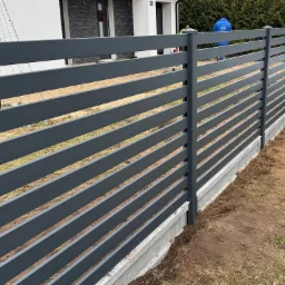 Nowoczesne, poziome, grafitowe przęsła ogrodzeniowe na betonowej podmurówce, widoczne z boku, z fragmentem elewacji domu w tle i pracownikiem w niebieskiej kurtce w oddali.