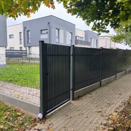 JAN-MET - Nowoczesne, grafitowe ogrodzenie panelowe z furtką, oddzielające posesję od chodnika. Widoczne domy w tle i opadłe liście. Oświetlenie w podstawie słupka.