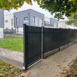 Nowoczesne, grafitowe ogrodzenie panelowe z furtką, oddzielające posesję od chodnika. Widoczne domy w tle i opadłe liście. Oświetlenie w podstawie słupka.