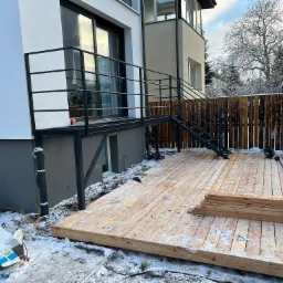Budowa drewnianego tarasu przy domu w zimowej scenerii, widoczne deski, częściowo ukończona podłoga i metalowe schody z balustradą.
