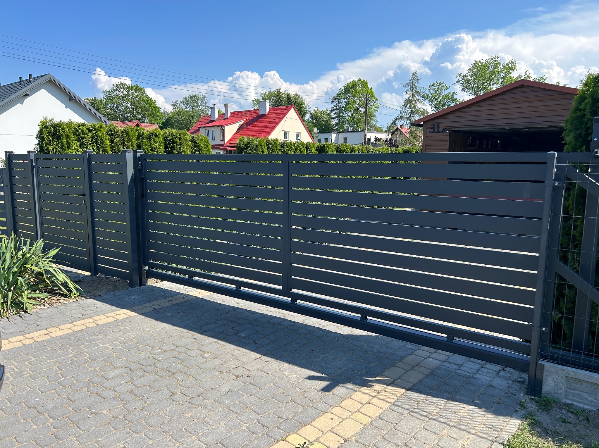 Nowoczesna, przesuwna brama wjazdowa z poziomych, grafitowych paneli na tle domów i błękitnego nieba z chmurami.