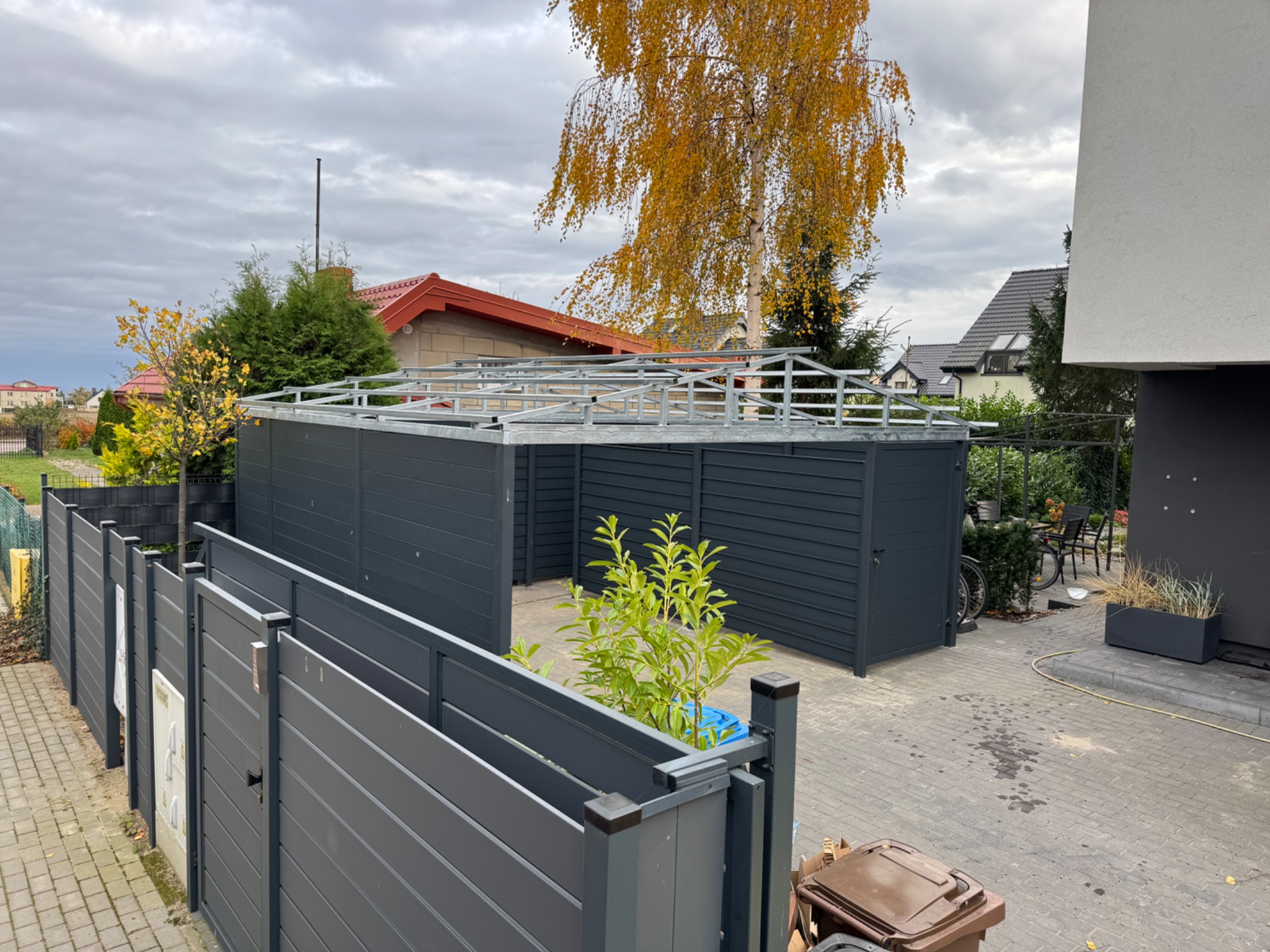 W trakcie budowy: szara konstrukcja z metalowym szkieletem dachu, prawdopodobnie wiata śmietnikowa, widoczna zza szarego ogrodzenia panelowego na posesji.