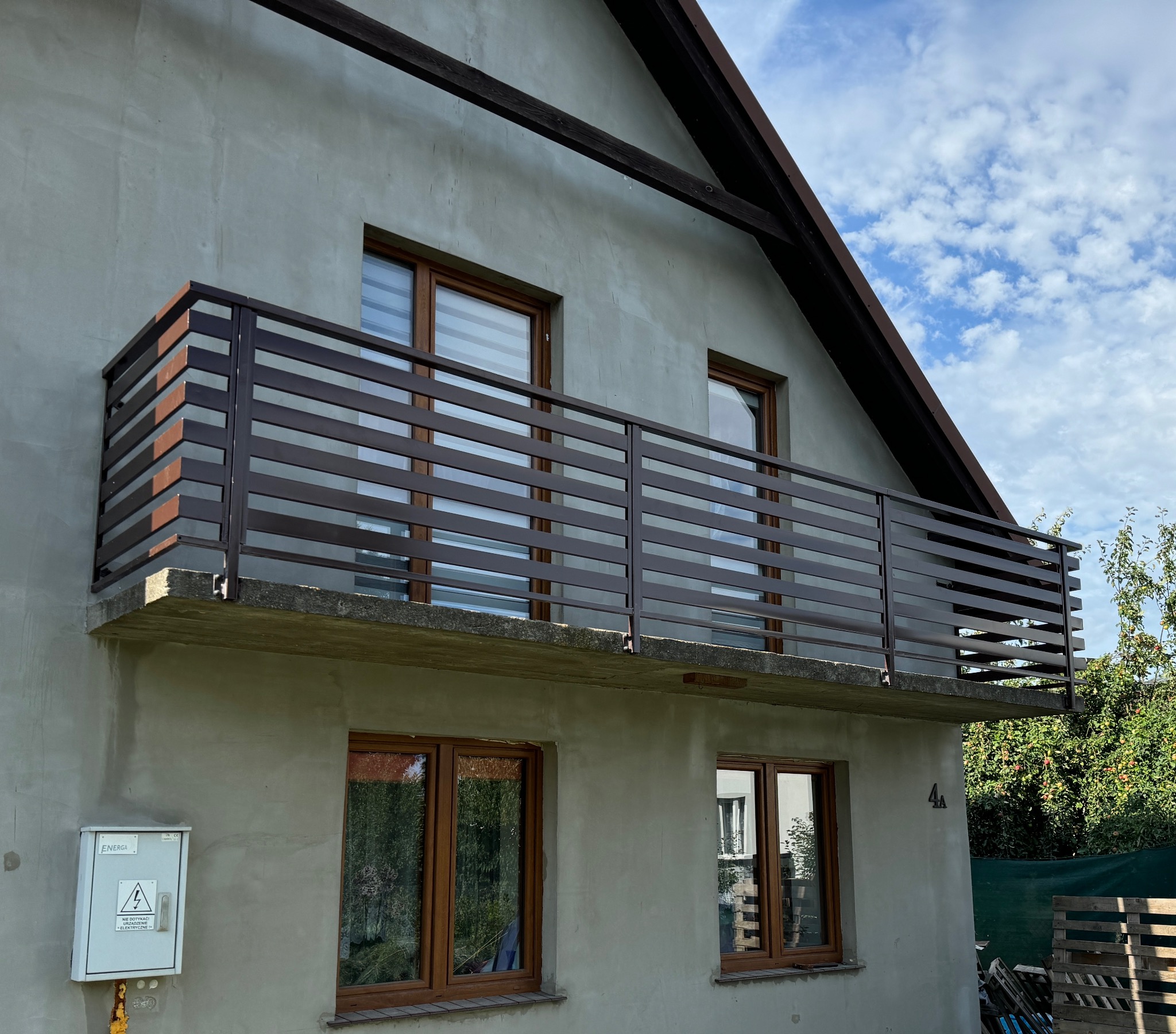 Nowoczesna, pozioma balustrada balkonowa z brązowych profili, zamontowana na betonowej płycie balkonu domu z surowym tynkiem i drewnianą stolarką okienną.