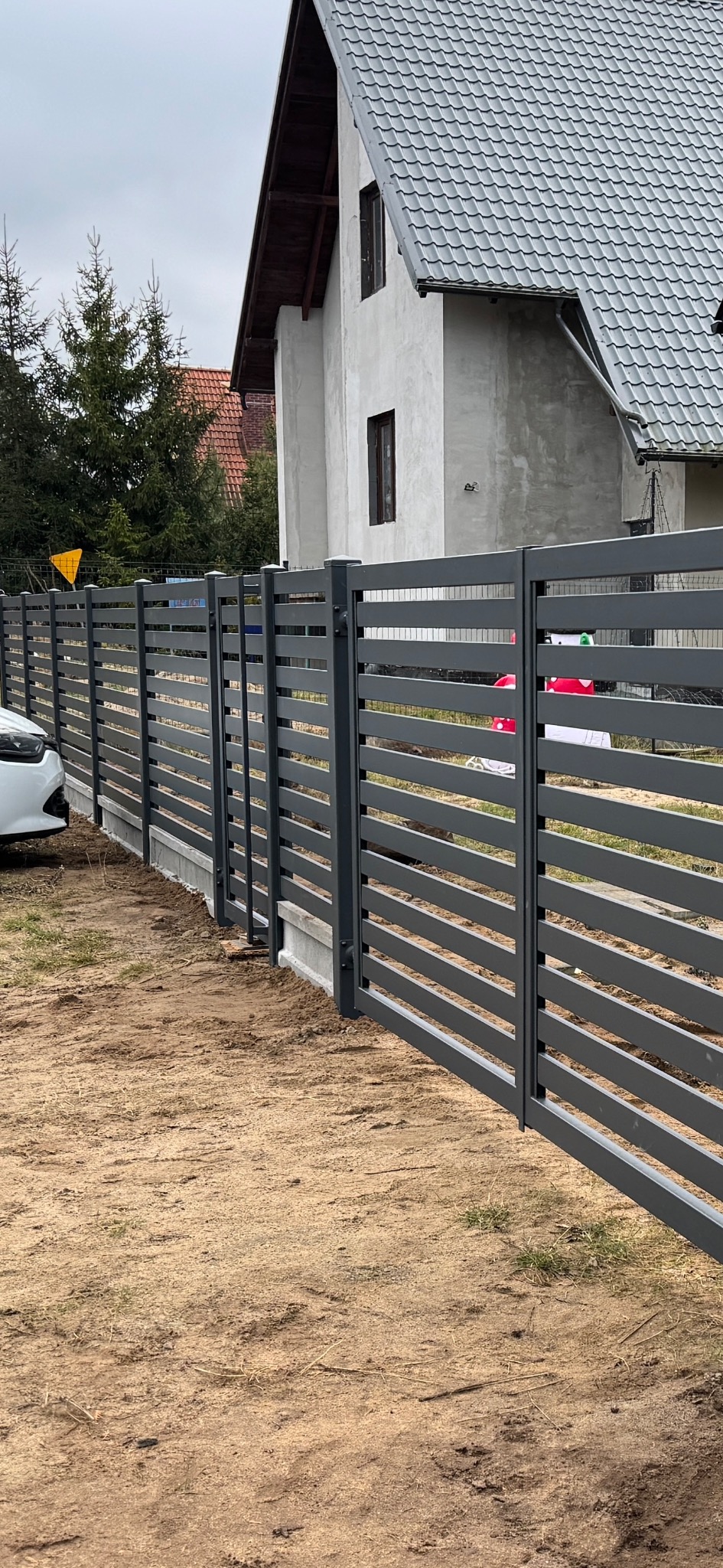 Nowoczesne, poziome przęsła w kolorze grafitowym tworzą ogrodzenie posesji z widocznym fragmentem białego samochodu i budynku mieszkalnego w tle. Widać betonową podmurówkę i surowy teren wokół.