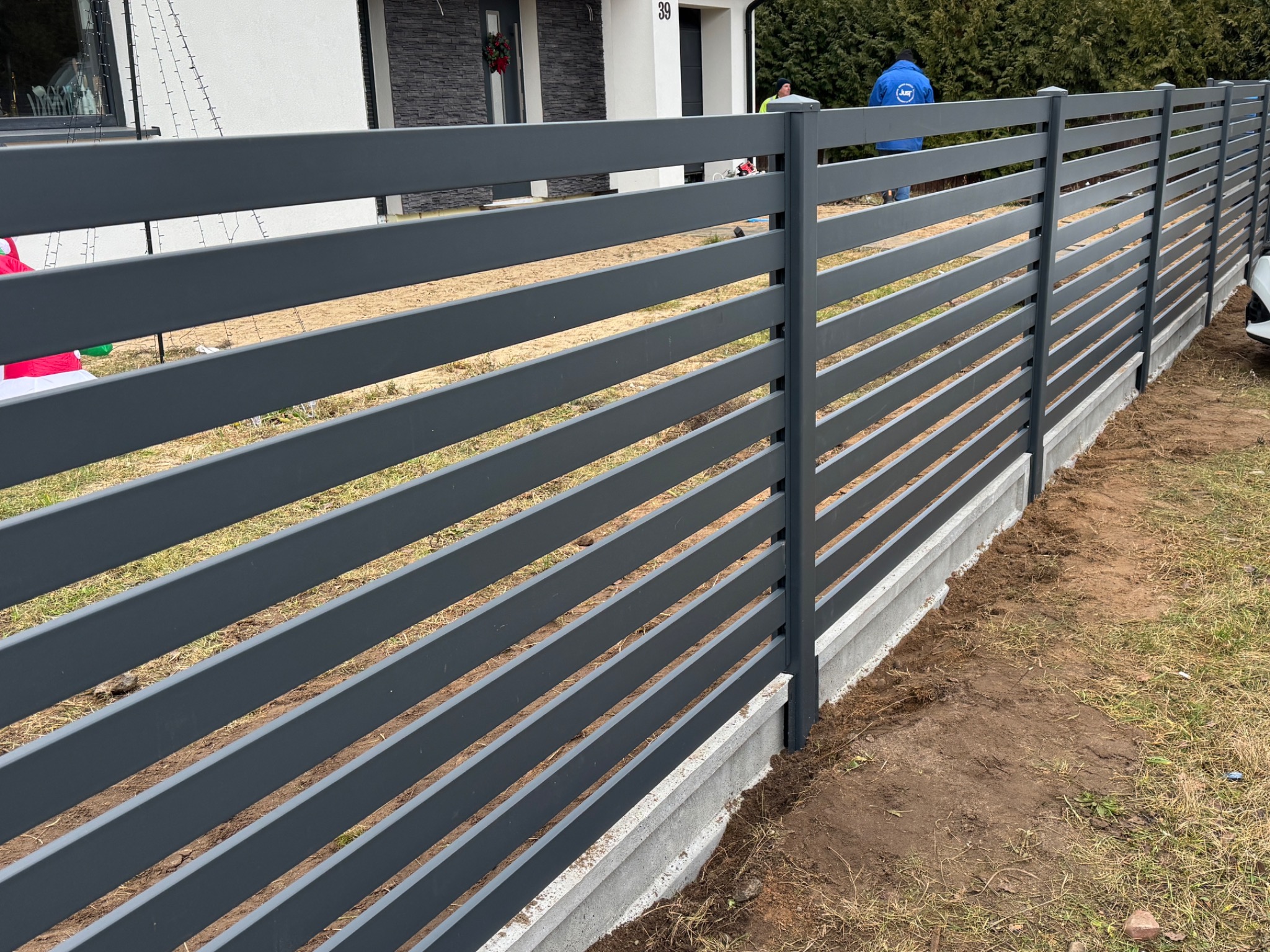 Nowoczesne, poziome, grafitowe przęsła ogrodzeniowe na betonowej podmurówce, widoczne z boku, z fragmentem elewacji domu w tle i pracownikiem w niebieskiej kurtce w oddali.