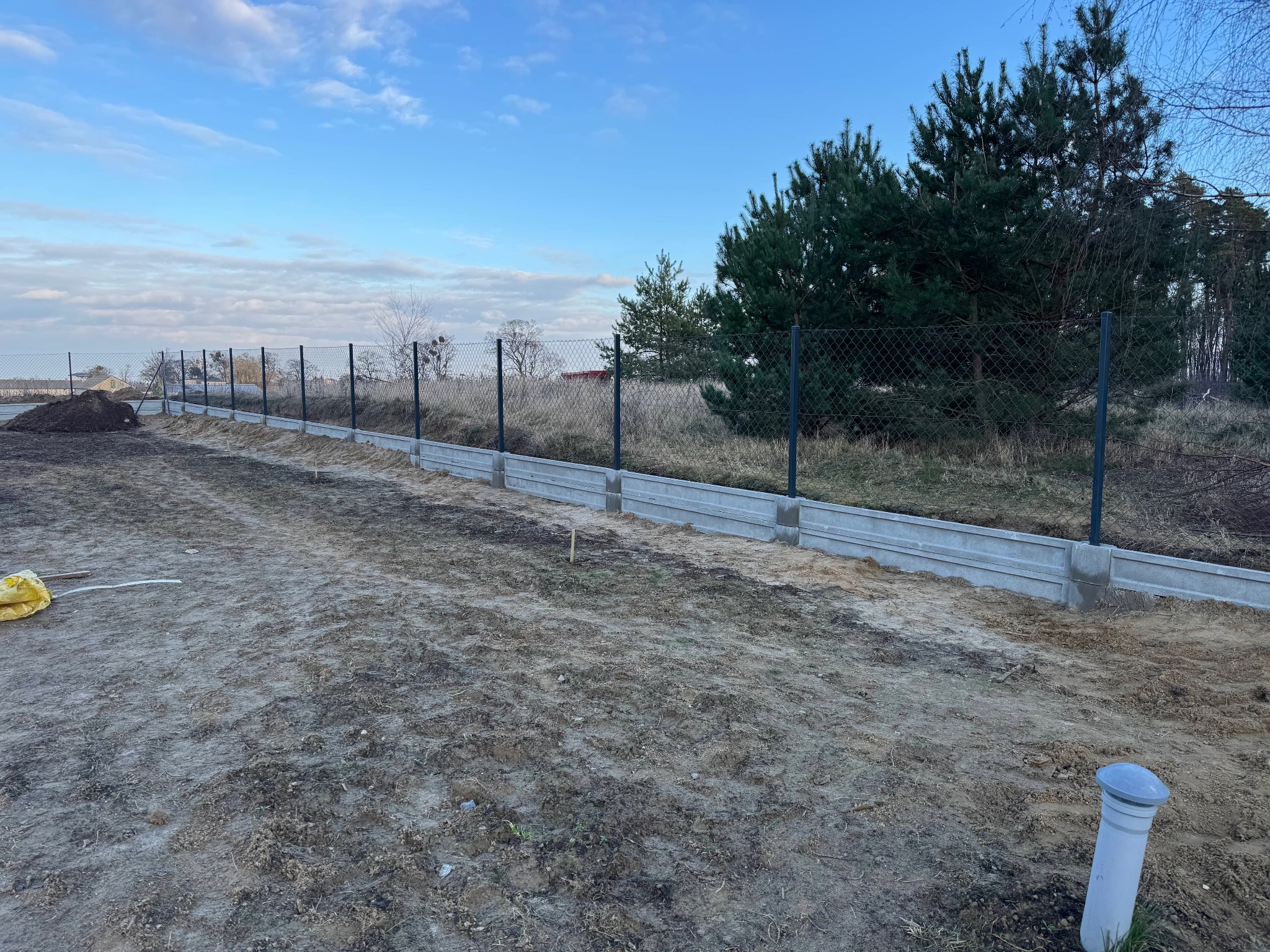Montaż siatki ogrodzeniowej na betonowej podmurówce, widoczny fragment terenu przed posesją z workiem i wystającą rurą wentylacyjną.