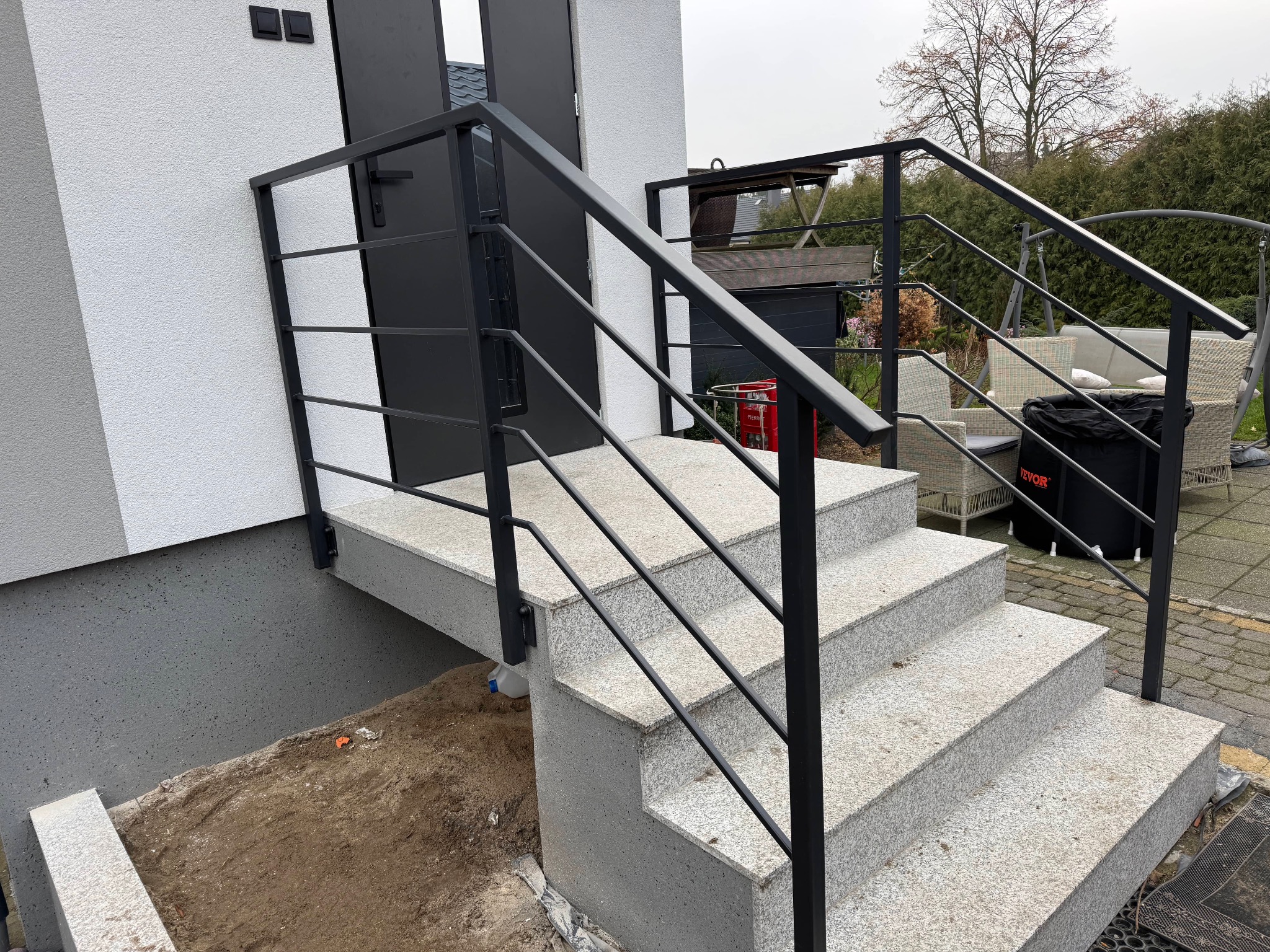 Zewnętrzne schody wejściowe z szarego granitu, zabezpieczone nowoczesną, czarną, metalową balustradą o minimalistycznym designie, widoczne z boku, z fragmentem elewacji budynku w tle.