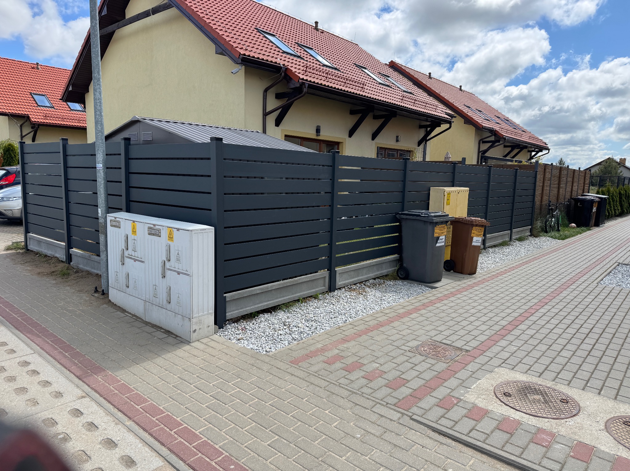 Nowoczesne ogrodzenie panelowe w kolorze grafitowym, oddzielające posesję od chodnika, z widoczną skrzynką elektryczną i koszami na śmieci. Tło stanowią domy z czerwoną dachówką.