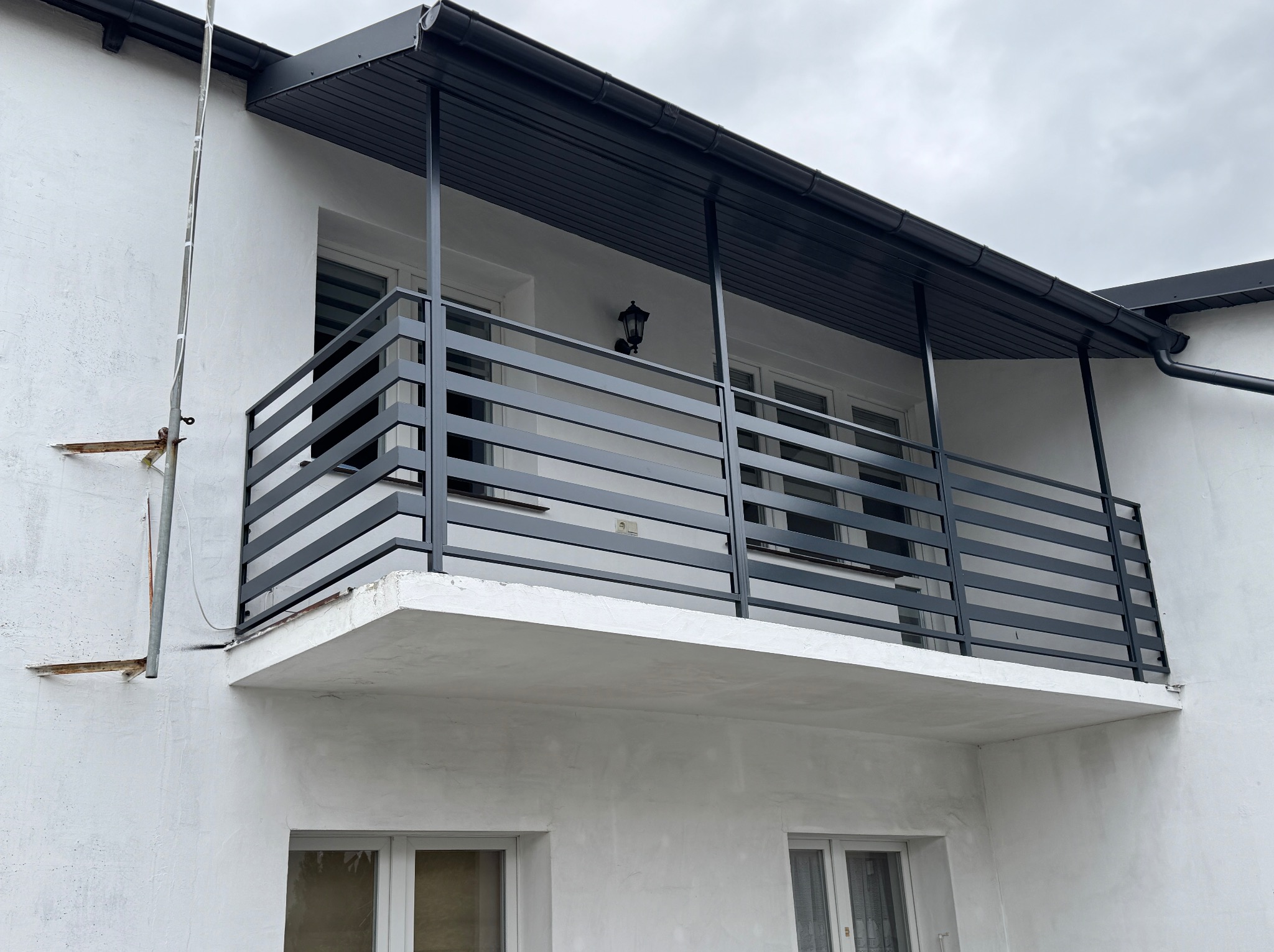 Nowoczesna, grafitowa balustrada balkonowa z poziomych profili, zamontowana na białym budynku z widocznymi oknami i zadaszeniem. Minimalistyczny design.