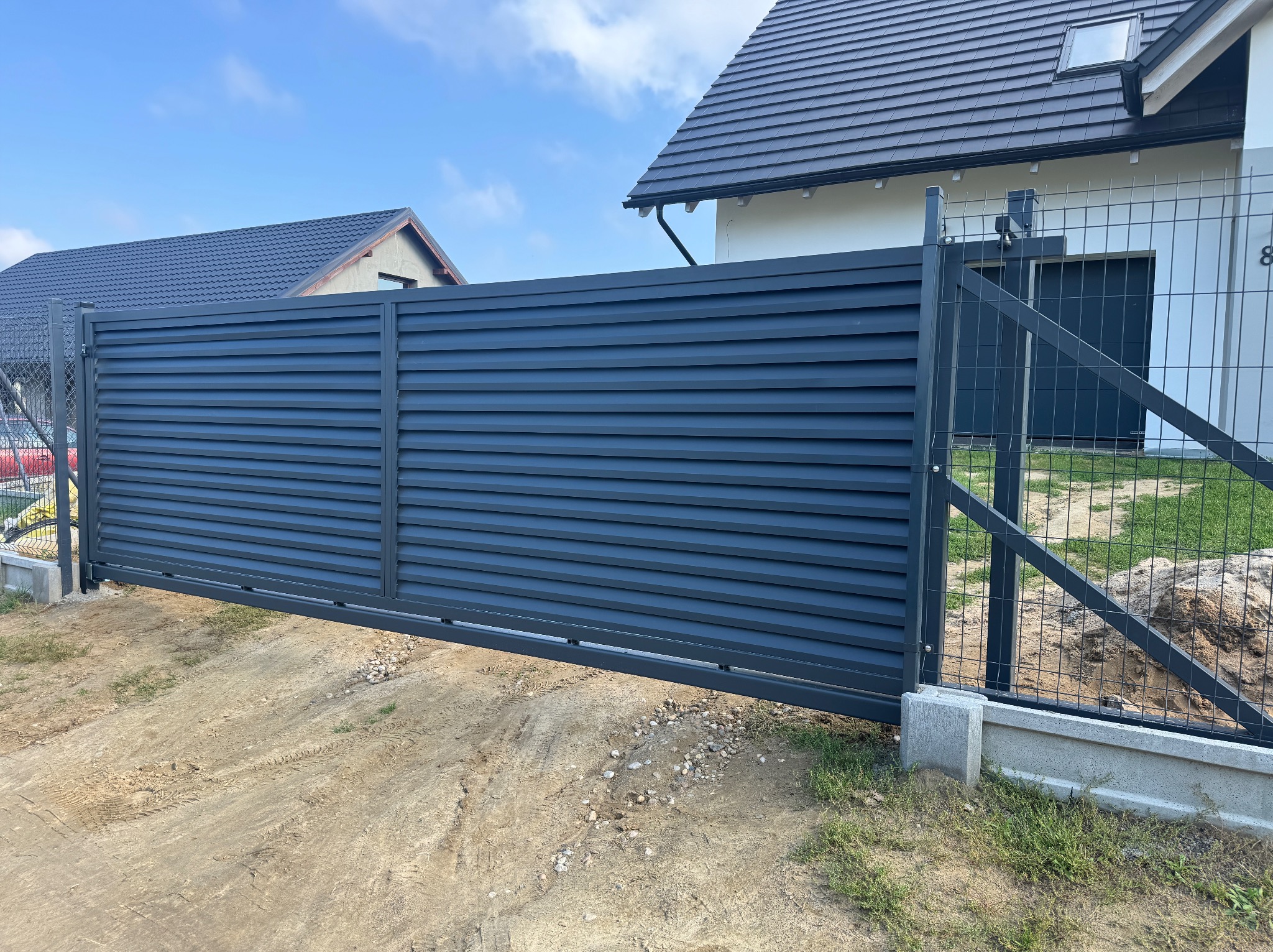 Nowoczesna przesuwna brama wjazdowa z poziomymi panelami w kolorze antracytowym, zamontowana przy domu z szarą dachówką i ogrodzeniem z siatki.