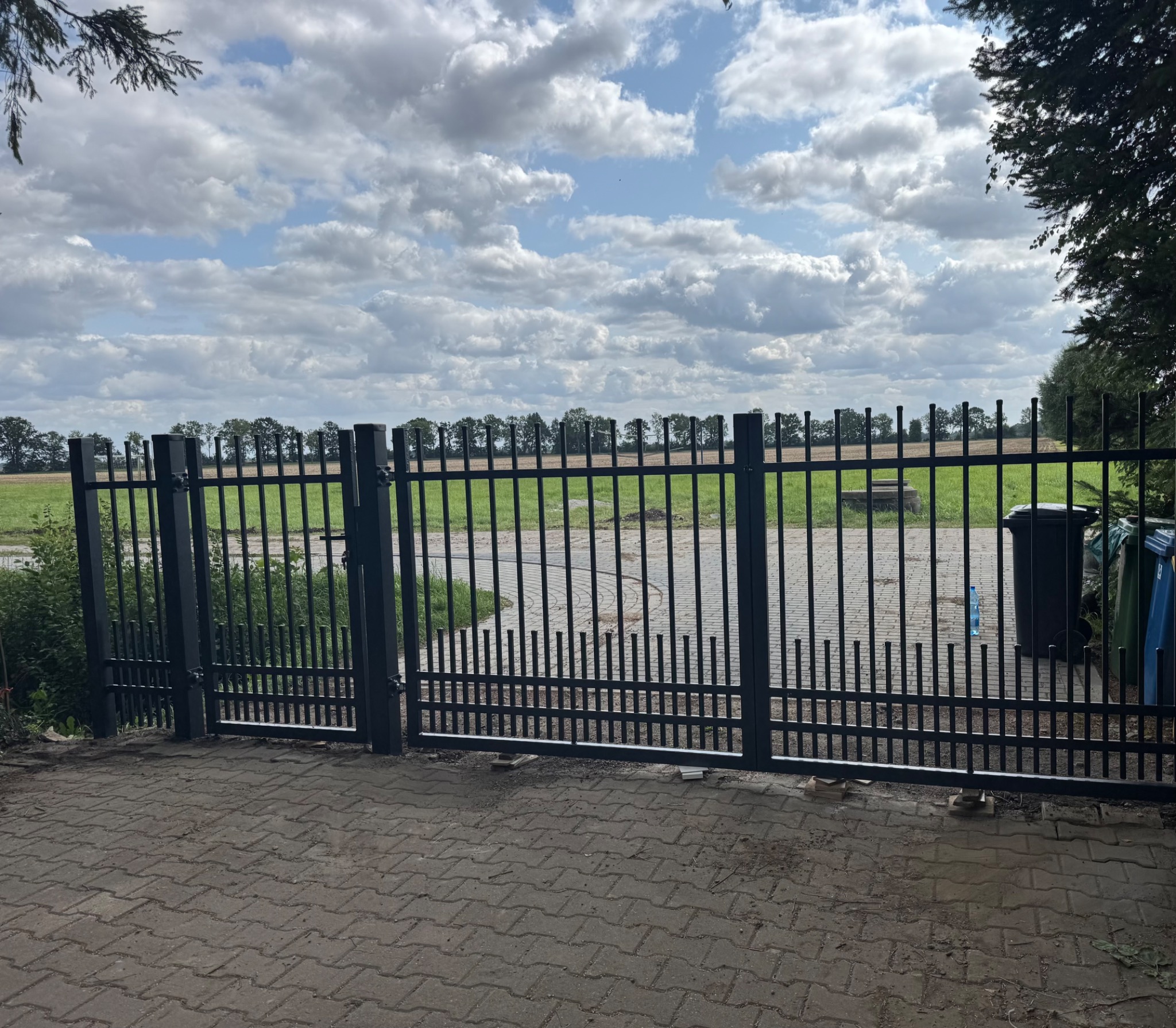 Nowoczesna brama wjazdowa z metalowymi sztachetami w Tczewie. Widok na polną przestrzeń przez bramę. Na pierwszym planie bruk, w tle drzewa i pochmurne niebo.