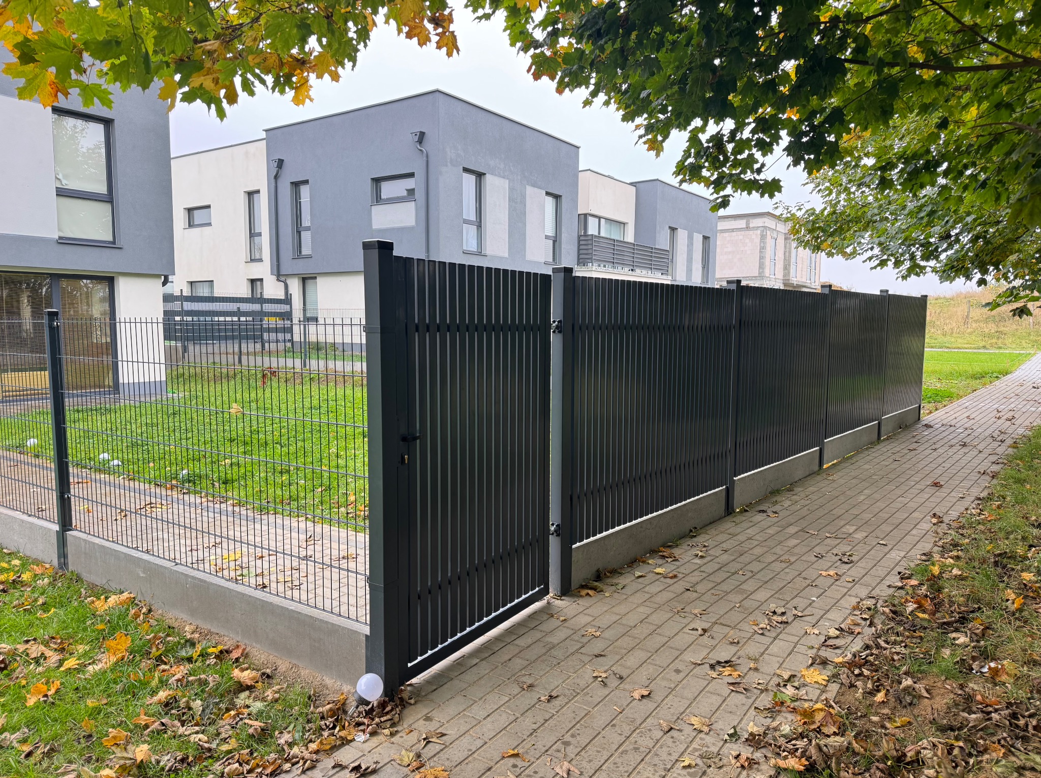 Nowoczesne, grafitowe ogrodzenie panelowe z furtką, oddzielające posesję od chodnika. Widoczne domy w tle i opadłe liście. Oświetlenie w podstawie słupka.