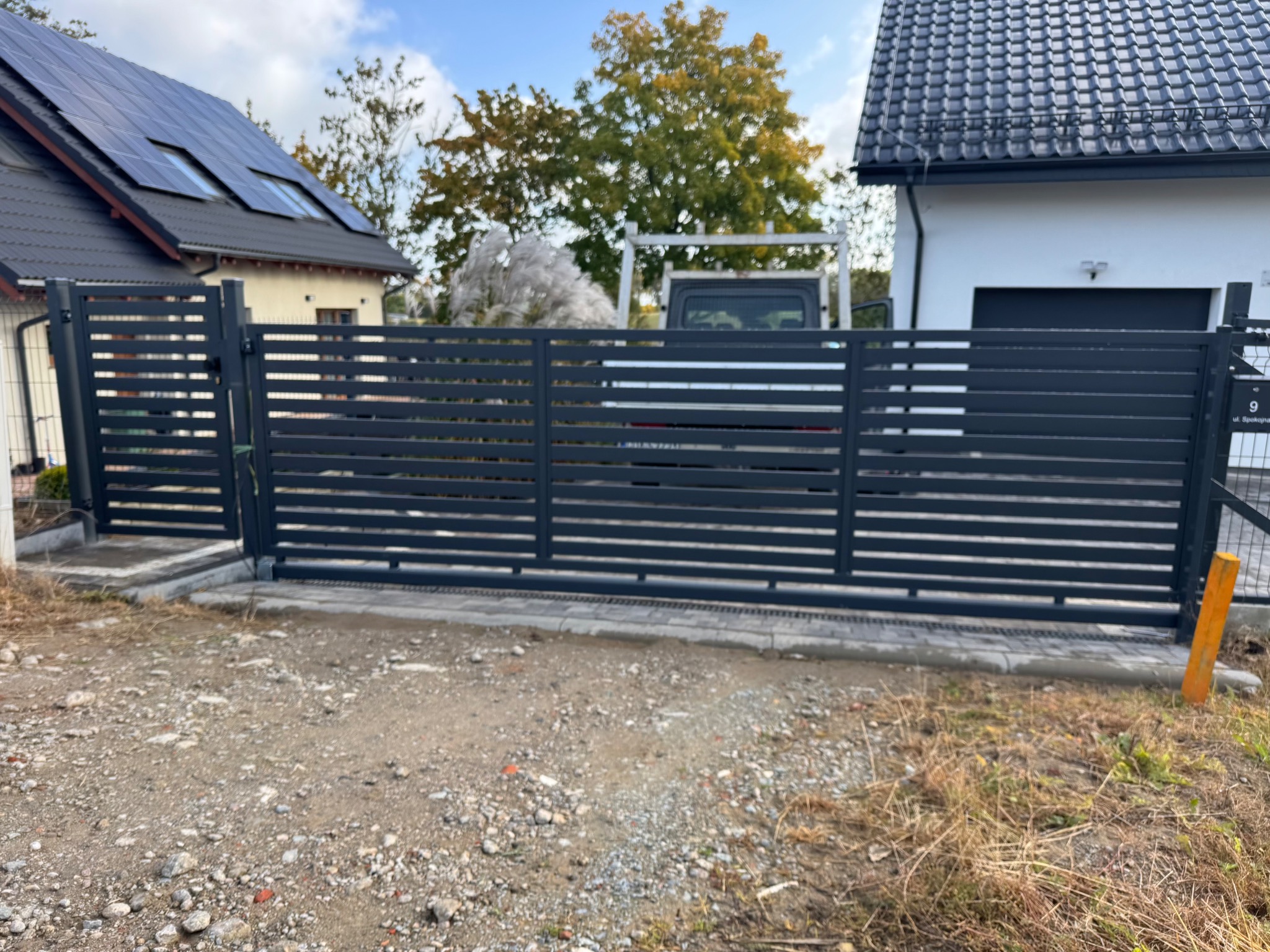 Nowoczesna, pozioma brama przesuwna w kolorze antracytowym, z widocznymi elementami montażowymi, na tle dwóch budynków. Widoczny fragment posesji.