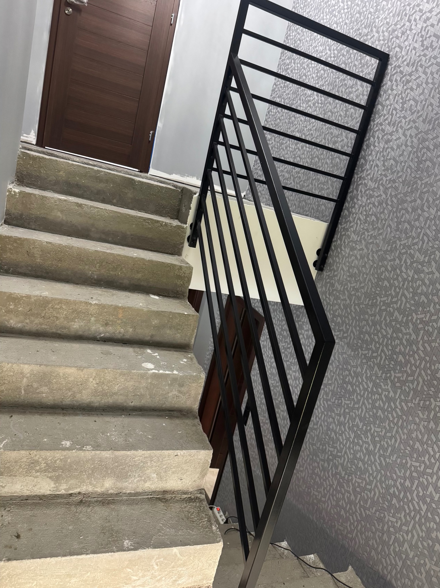 Czarne, metalowe poręcze schodowe o prostej konstrukcji, zamontowane na surowych, betonowych schodach. W tle widoczna ściana z geometrycznym wzorem i drewniane drzwi.