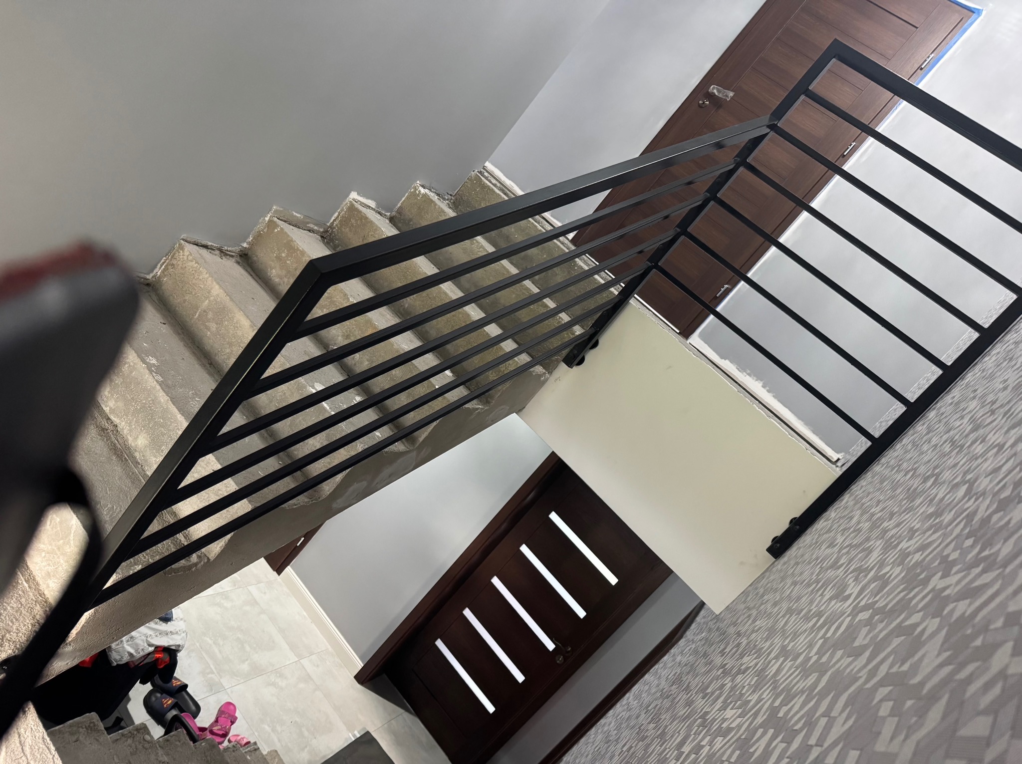 Minimalistyczna, czarna poręcz schodowa z poziomymi szczeblami, zamontowana na surowych, betonowych schodach. Widok z góry, drzwi w tle.