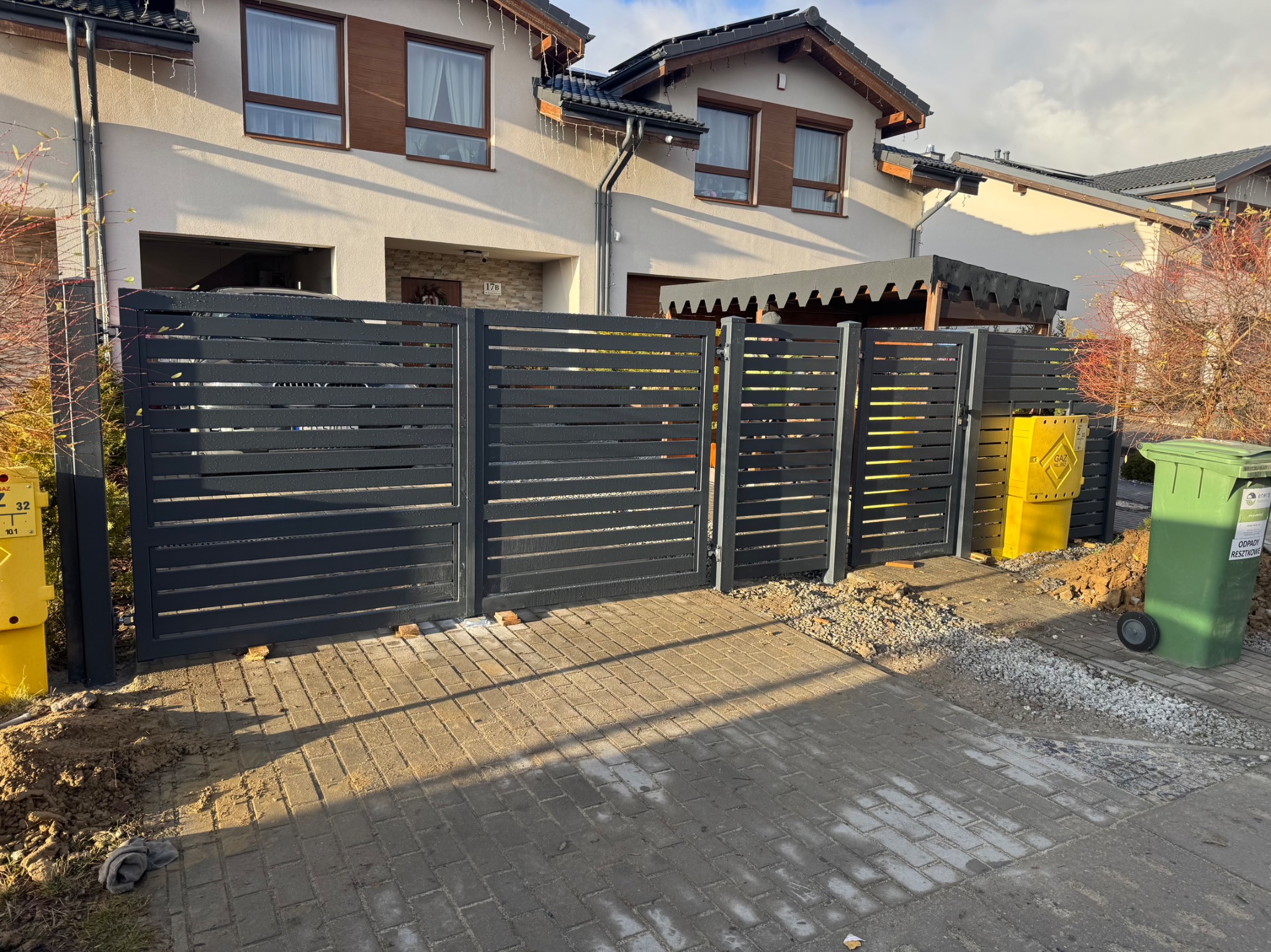 Nowoczesna, pozioma brama wjazdowa z metalowych profili w kolorze grafitowym, na tle domu jednorodzinnego z jasną elewacją i brązowymi oknami. Widoczne kosze na śmieci.