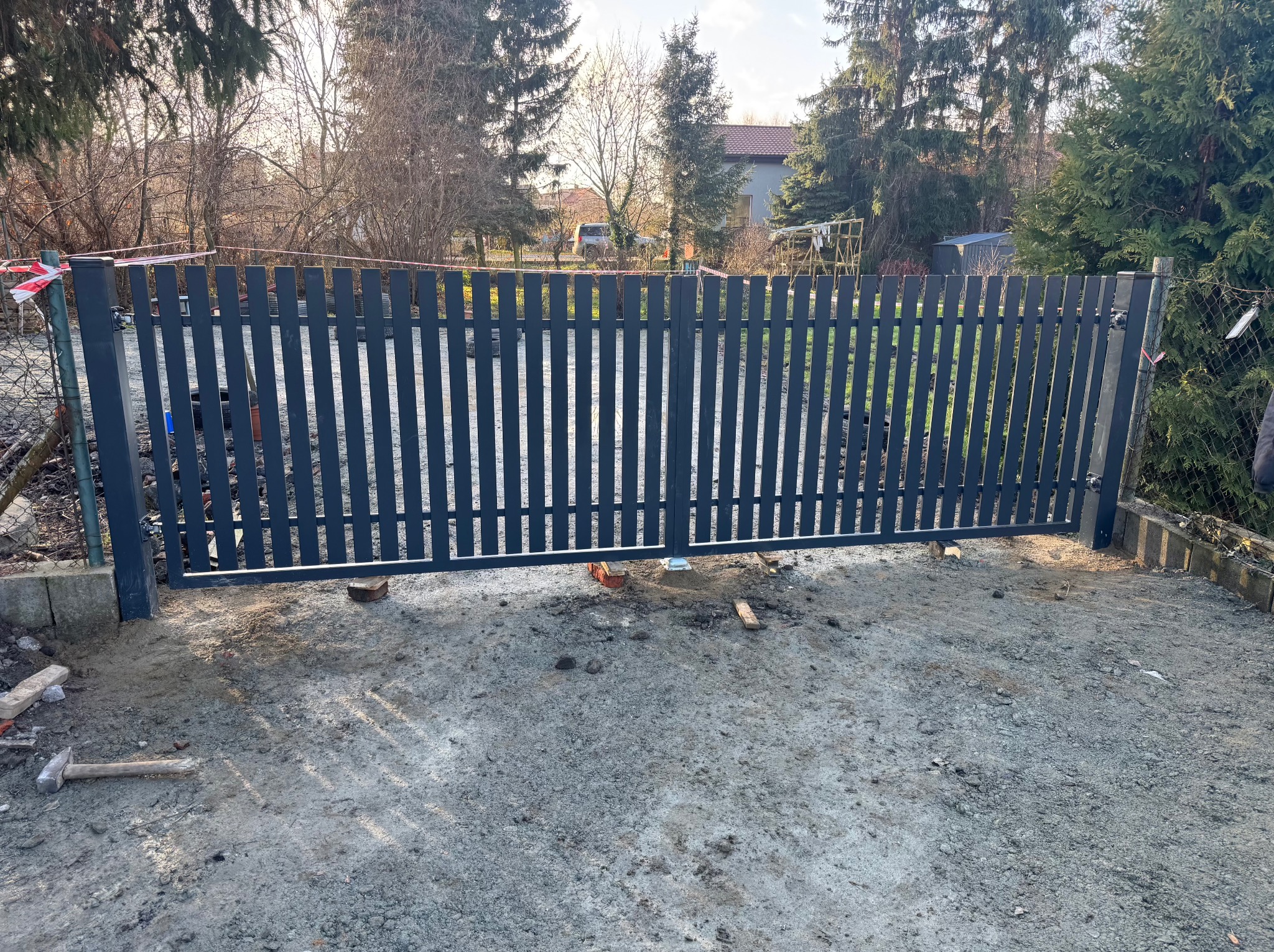 Nowoczesna, grafitowa brama wjazdowa z pionowymi szczebelkami, częściowo zamontowana na posesji. Widoczne narzędzia i elementy montażowe. Tło: drzewa i budynek.