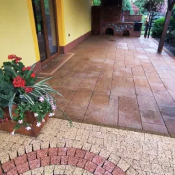 Zadaszony taras z brązowej kostki brukowej, drewniane donice z czerwonymi i białymi kwiatami, murowany grill w tle, żółta ściana domu.