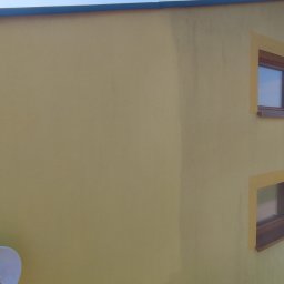 HI-LIFT Patryk Bieniaszewski - Żółta elewacja budynku z dwoma oknami w brązowych ramach, fragmentem elewacji w innym odcieniu żółtego i anteną satelitarną, widoczne ślady zabrudzeń na ścianie.