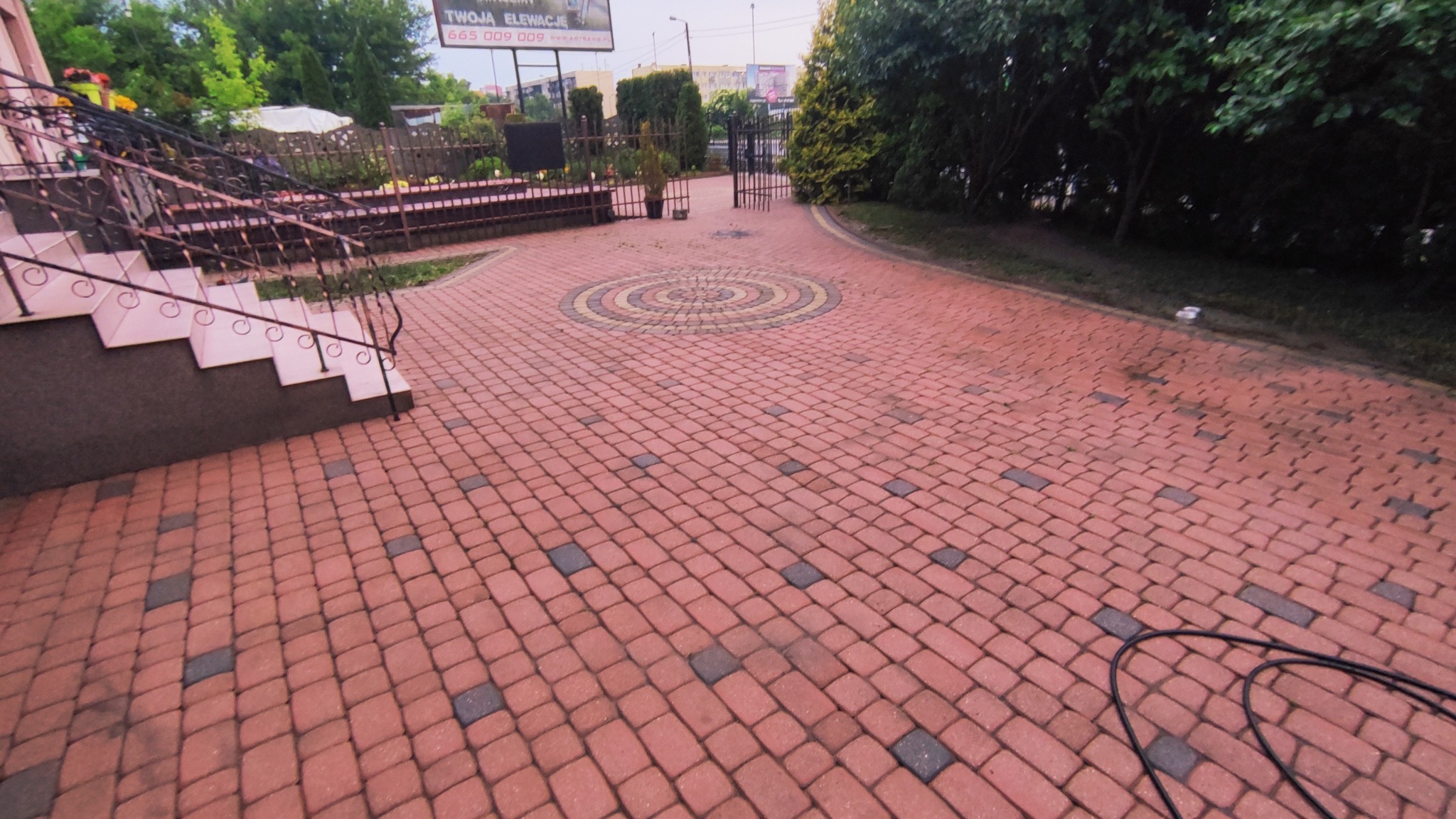 Ułożona z czerwonej kostki brukowej powierzchnia podjazdu z dekoracyjnym okręgiem z kostki w centrum, widoczne schody i zieleń w tle.