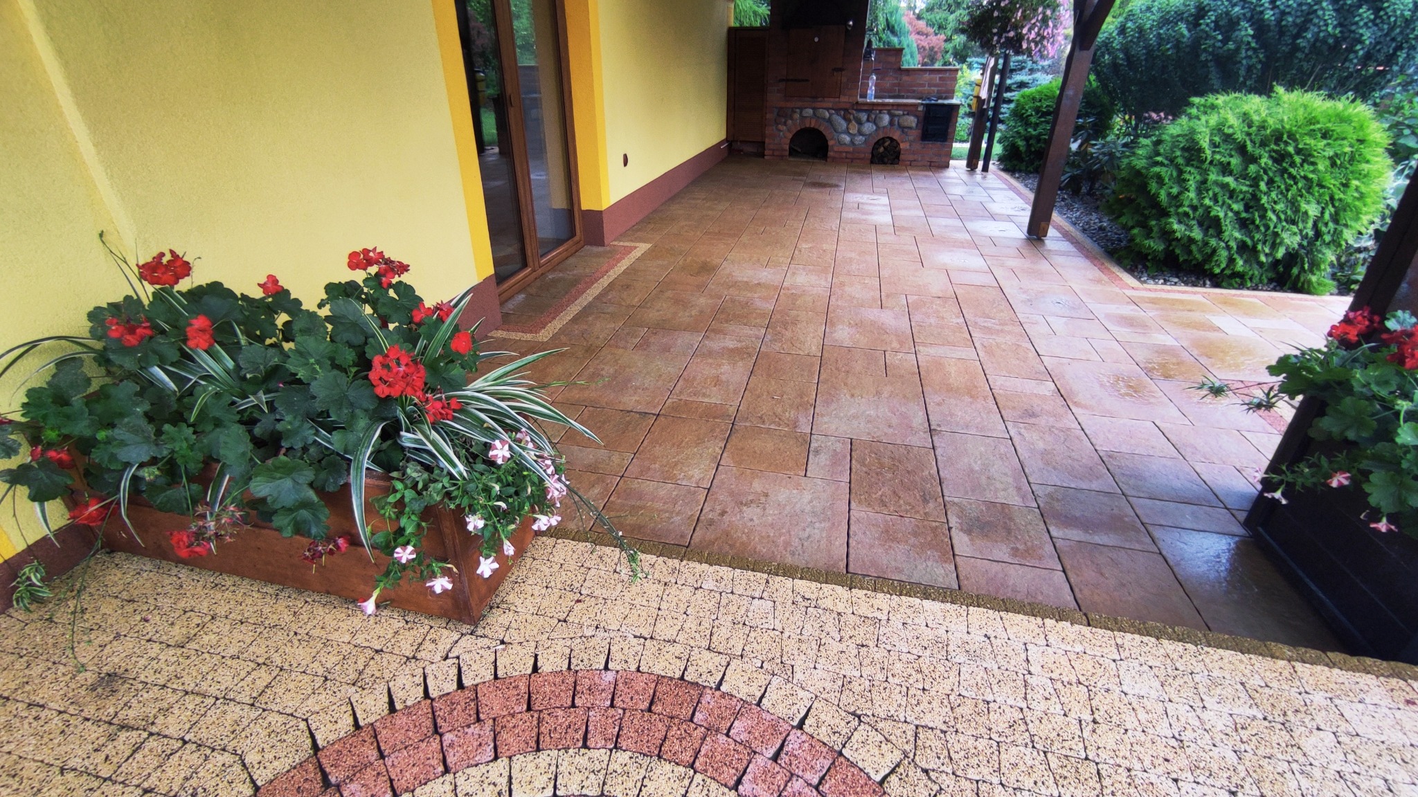 Zadaszony taras z brązowej kostki brukowej, drewniane donice z czerwonymi i białymi kwiatami, murowany grill w tle, żółta ściana domu.