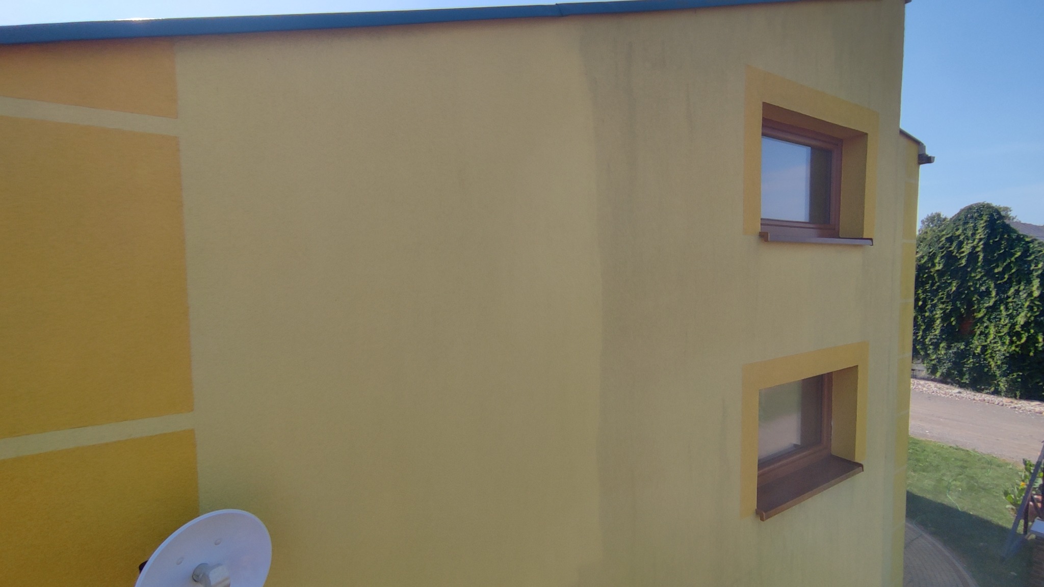 Żółta elewacja budynku z dwoma oknami w brązowych ramach, fragmentem elewacji w innym odcieniu żółtego i anteną satelitarną, widoczne ślady zabrudzeń na ścianie.