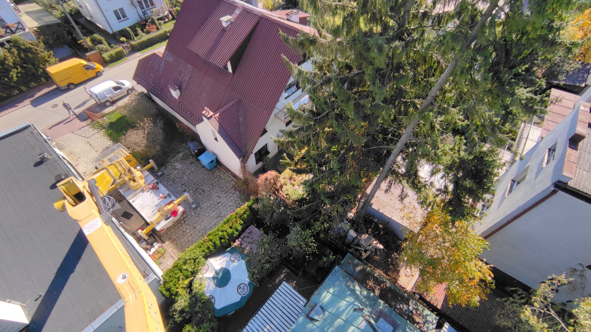 Widok z góry na żółty podnośnik koszowy w Lublinie, zaparkowany obok domu z czerwonym dachem, z widocznym drzewem i fragmentem ogrodu.