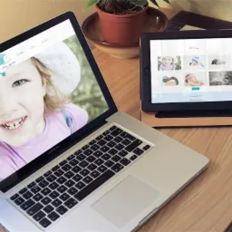 Laptop i tablet wyświetlające stronę internetową z dużym zdjęciem uśmiechniętej dziewczynki w kapeluszu, obok doniczka z rośliną na drewnianym biurku.