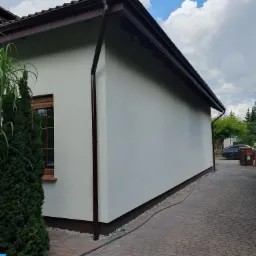 Świeżo otynkowana ściana budynku jednorodzinnego w Poznaniu, z brązowymi rynnami i okapem, widoczna z poziomu brukowanego podjazdu.