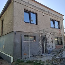 TOM-MAT Mateusz Mamot - Dwupiętrowy dom w trakcie remontu elewacji, z widocznymi nowymi oknami i fragmentami starej ściany za metalowym ogrodzeniem. Część ściany ocieplona.