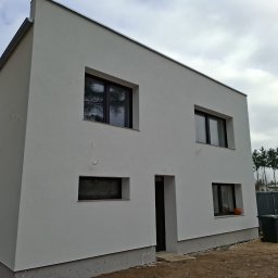 TOM-MAT Mateusz Mamot - Nowoczesny, dwukondygnacyjny dom z białą elewacją i ciemnymi oknami. Na parapecie pomarańczowa dynia. Widoczne drzewa w odbiciu okiennym i szary kosz na śmieci.