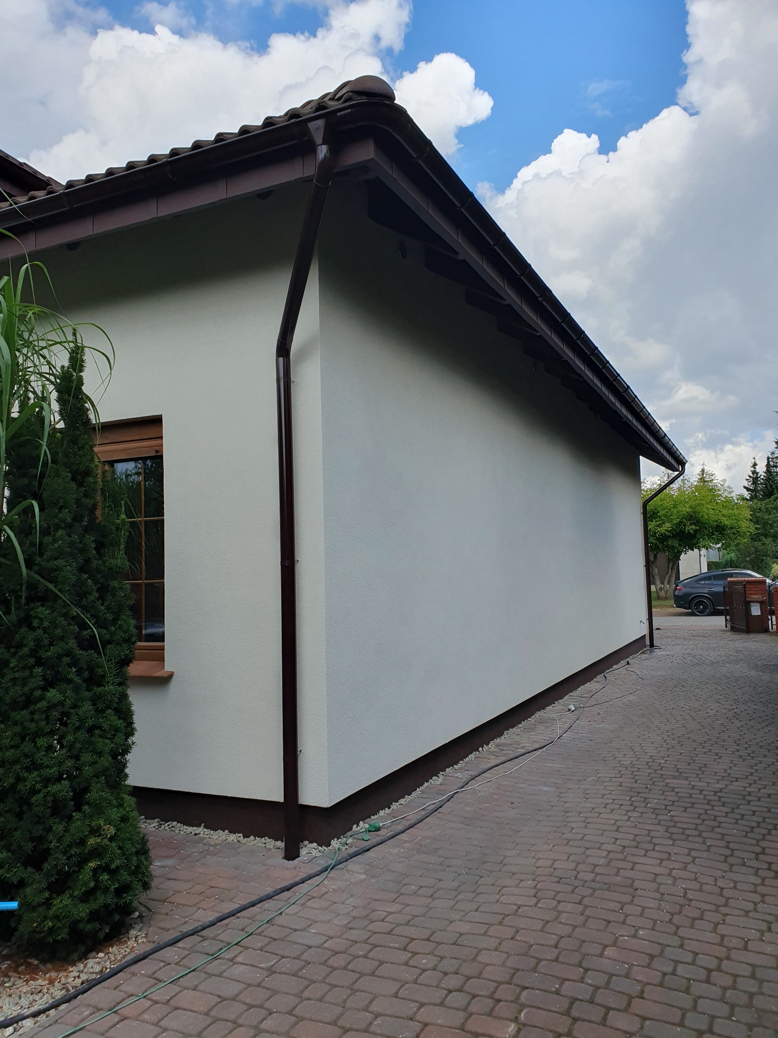 Świeżo otynkowana ściana budynku jednorodzinnego w Poznaniu, z brązowymi rynnami i okapem, widoczna z poziomu brukowanego podjazdu.