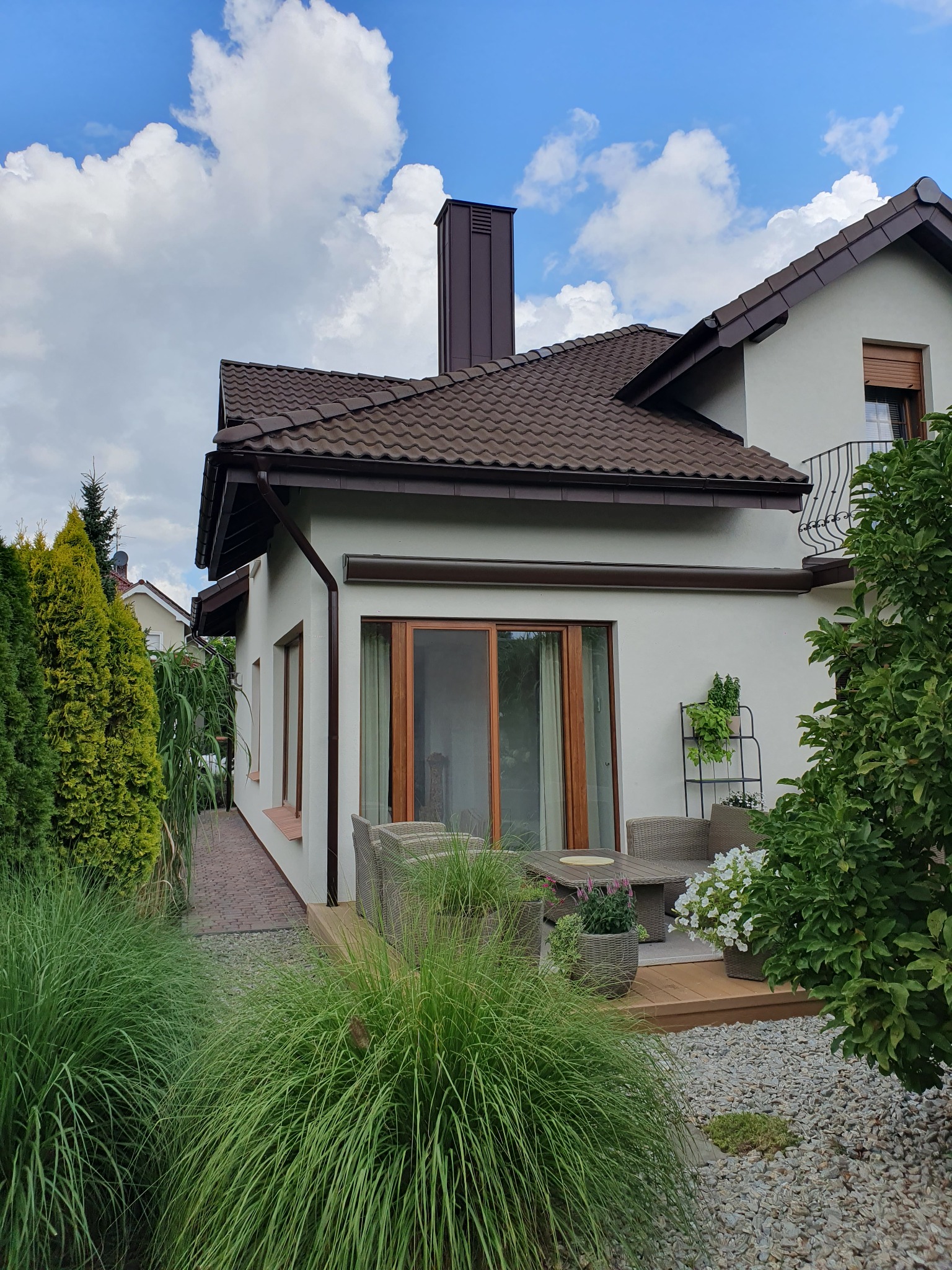 Jasny dom jednorodzinny z brązowym dachem i kominem, widok z ogrodu z ozdobnymi trawami i kamieniami, drewniany taras z meblami ogrodowymi.