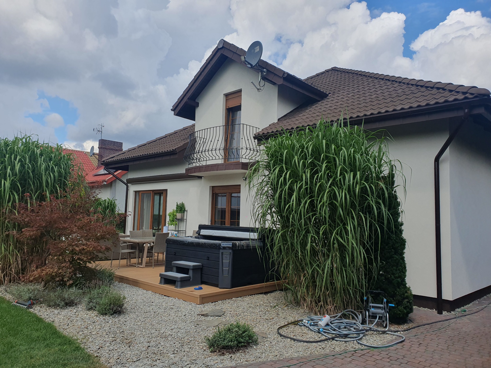 Dwupiętrowy dom z balkonem, tarasem z jacuzzi i zadbanym ogrodem z ozdobnymi trawami i kamiennym podłożem.