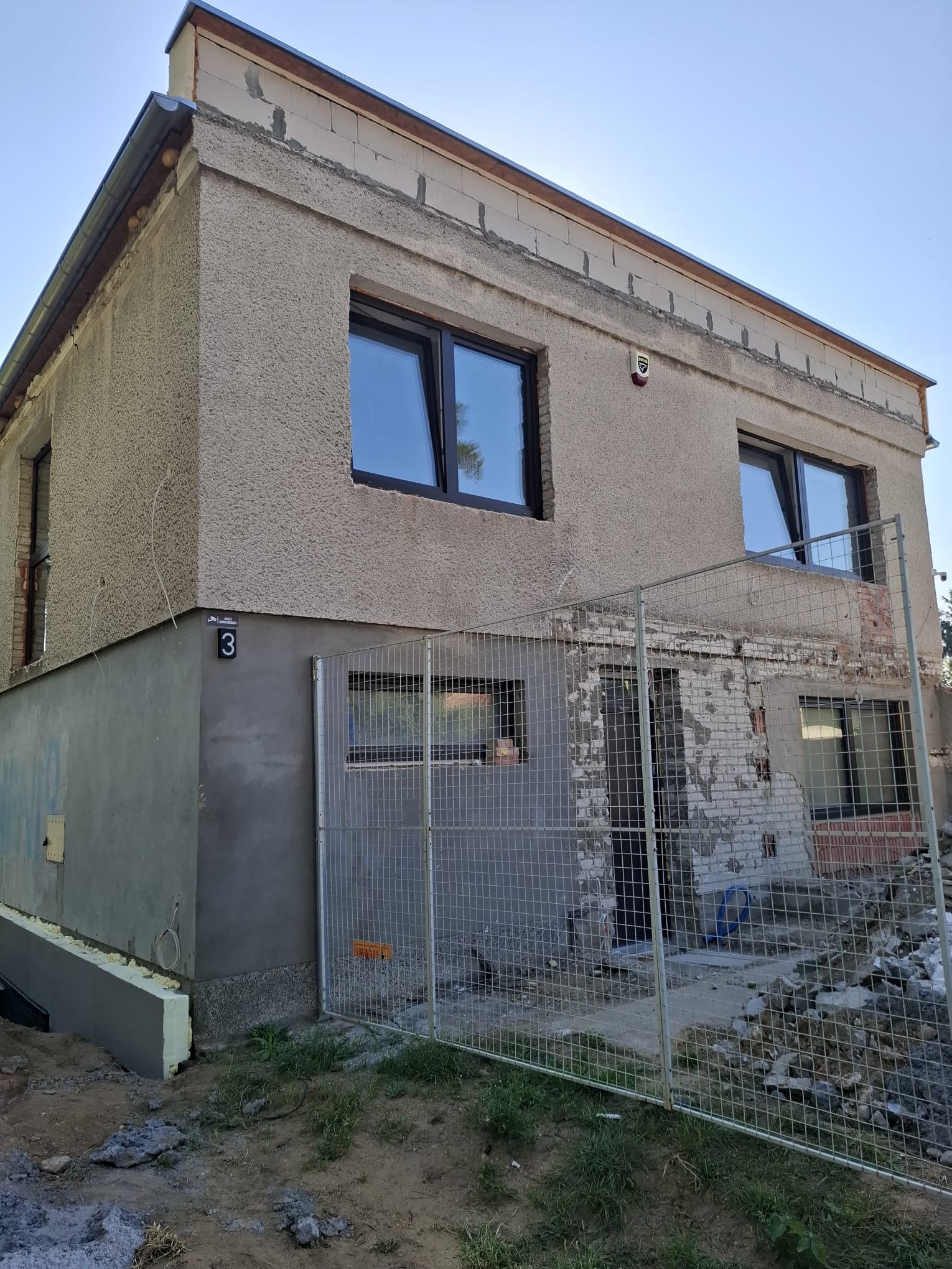 Dwupiętrowy dom w trakcie remontu elewacji, z widocznymi nowymi oknami i fragmentami starej ściany za metalowym ogrodzeniem. Część ściany ocieplona.