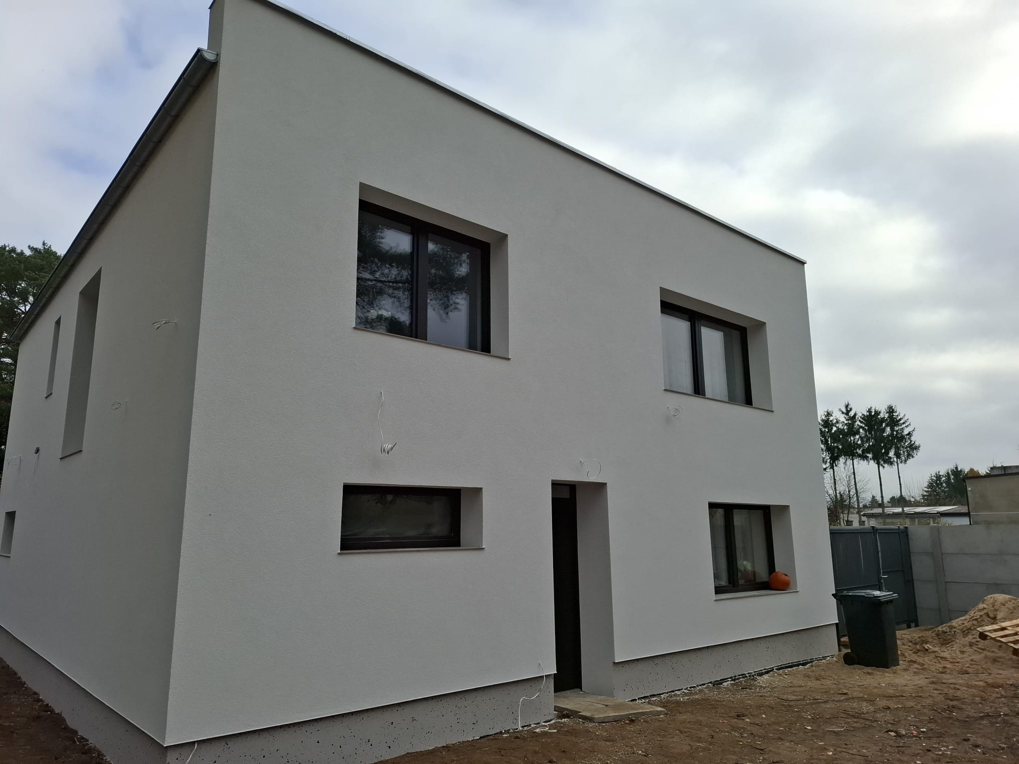 Nowoczesny, dwukondygnacyjny dom z białą elewacją i ciemnymi oknami. Na parapecie pomarańczowa dynia. Widoczne drzewa w odbiciu okiennym i szary kosz na śmieci.