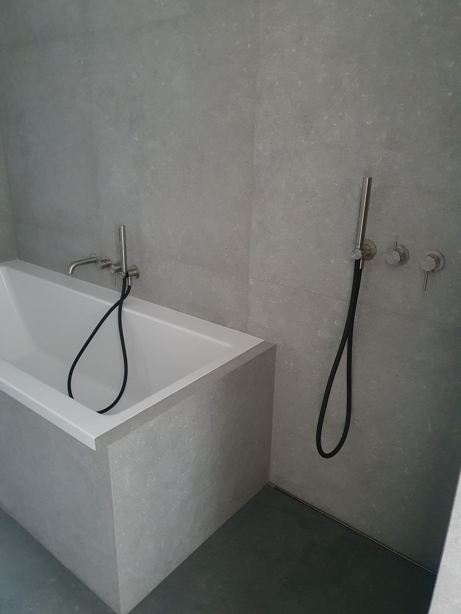 Minimalistyczna łazienka z wanną obłożoną szarą płytką, bateria wannowa i prysznic ręczny w kolorze srebrnym, szare płytki na ścianach i podłodze.