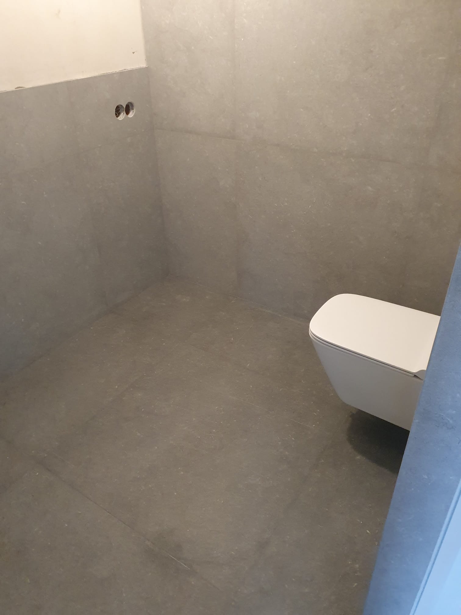 Wykończona łazienka z szarymi płytkami i podwieszaną toaletą. Widoczne otwory na armaturę. Minimalistyczny design.