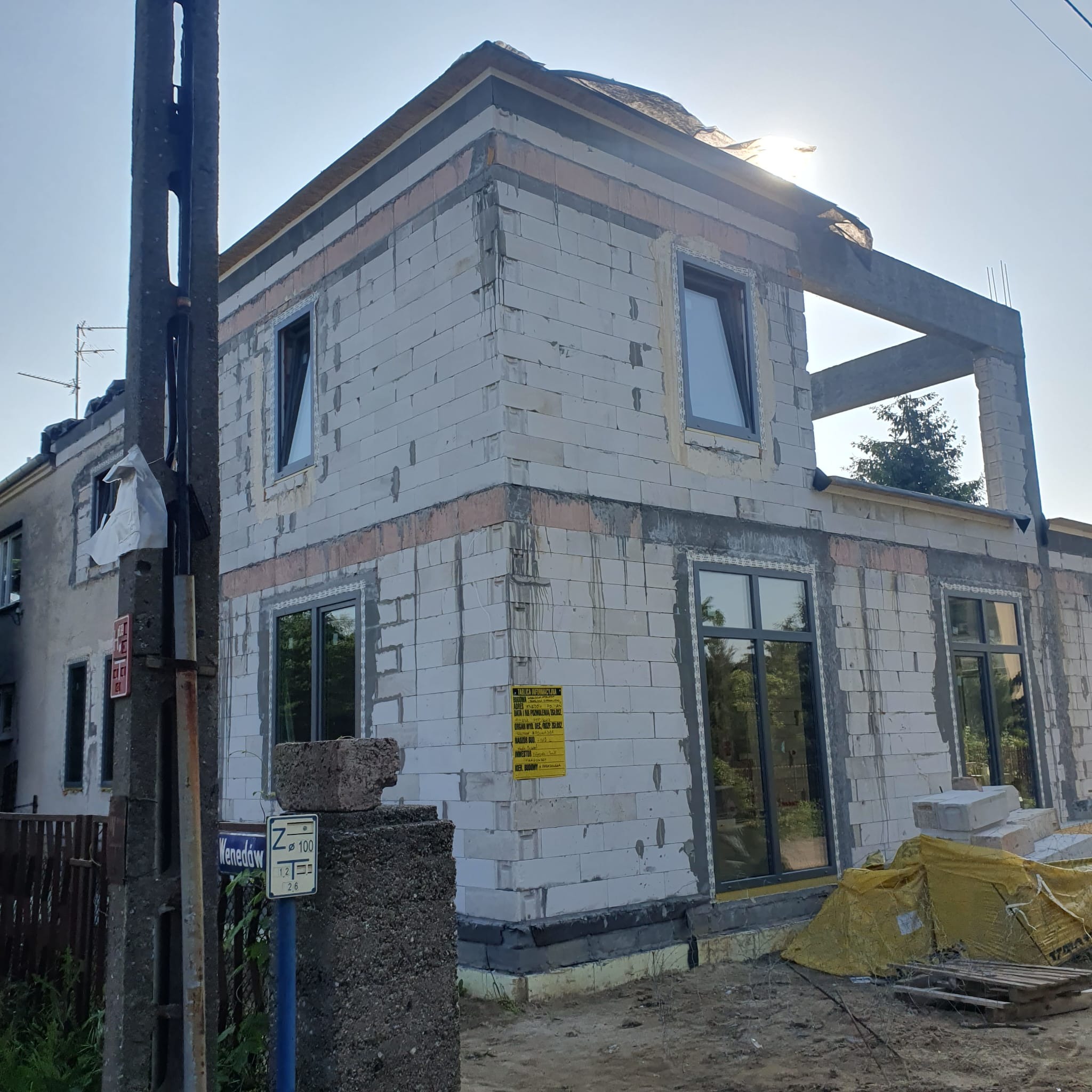 Surowy stan domu w trakcie rozbudowy, widoczne białe bloczki, okna z ciemnymi ramami, betonowe nadproża i elementy konstrukcyjne. W tle słoneczne niebo.