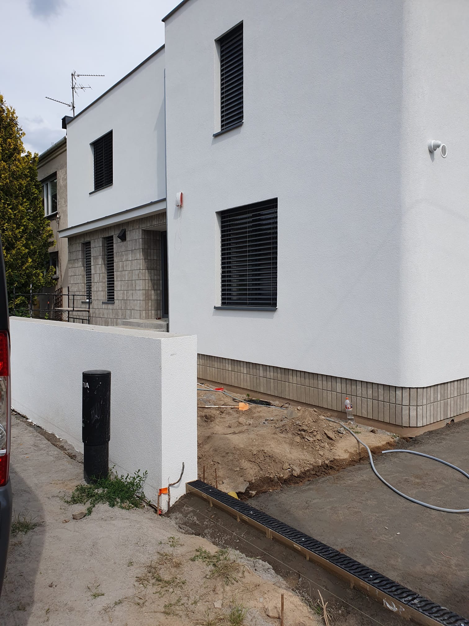 Biały, dwukondygnacyjny dom z płaskim dachem i ciemnymi żaluzjami w oknach. Widoczny fragment elewacji z wykończeniem z płytek. Teren wokół domu w trakcie prac wykończeniowych.