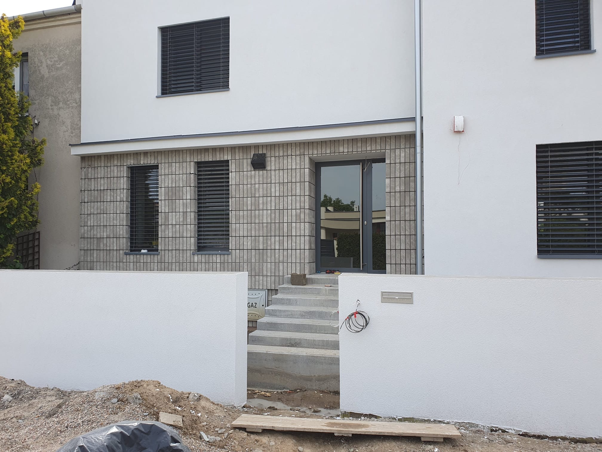 Nowoczesny dom z jasną elewacją i elementami z cegły klinkierowej. Widoczne schody wejściowe, okna z żaluzjami zewnętrznymi i fragment ogrodu. Minimalistyczny design.