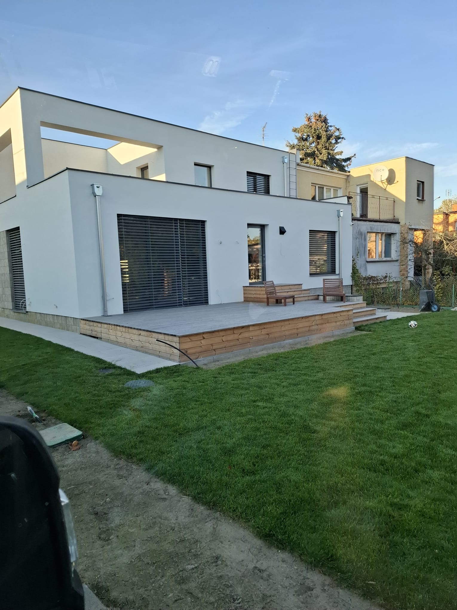 Nowoczesny dom z białymi ścianami i drewnianym tarasem. Widoczne detale architektoniczne i zadbany trawnik. Ujęcie z poziomu trawnika, w tle inne budynki.