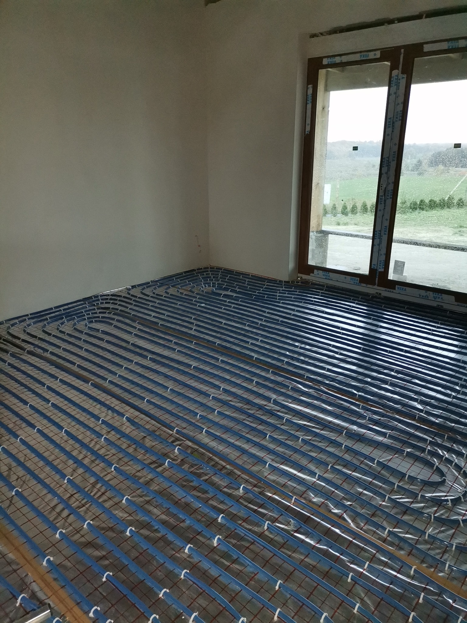 Niebieskie rurki ogrzewania podłogowego ułożone na folii izolacyjnej w surowym pomieszczeniu z oknem.