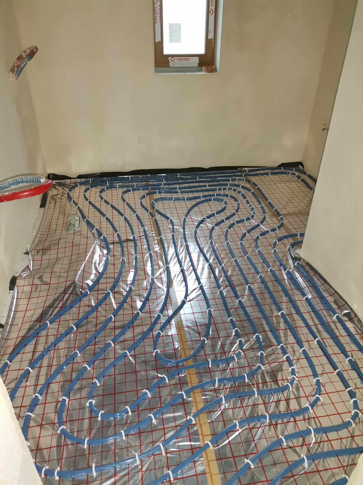 Niebieskie rury ogrzewania podłogowego ułożone na folii z czerwoną siatką, widok z góry na niedokończoną instalację w jasnym pomieszczeniu.