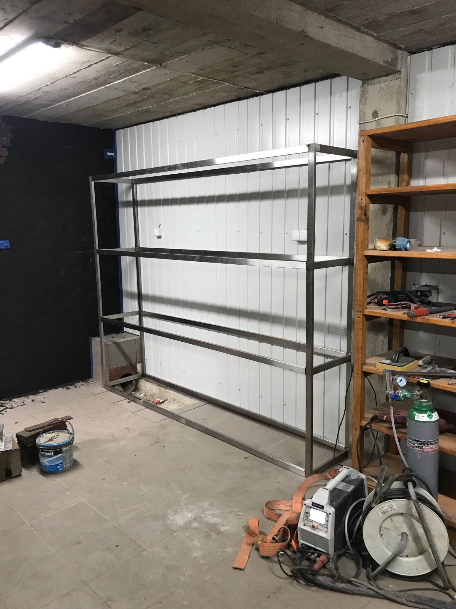 Surowa stalowa konstrukcja regału z trzema półkami w trakcie budowy w pomieszczeniu z surowym betonowym sufitem, białymi panelami ściennymi i widocznymi narzędziami do obróbki metali.