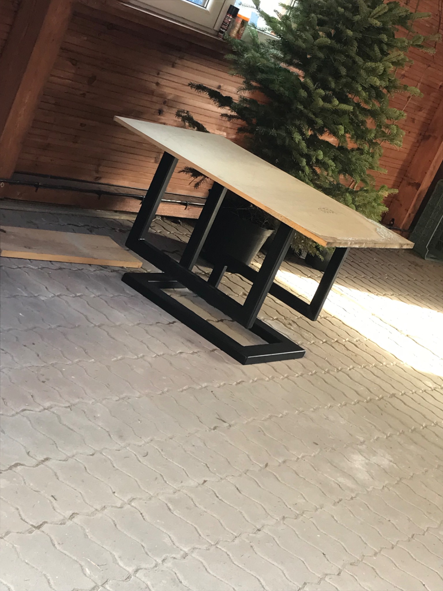 Stół z drewnianym blatem i czarną, metalową podstawą o geometrycznym kształcie, umieszczony na betonowej kostce brukowej, z widocznym fragmentem drewnianej ściany i zielonego drzewka w tle.