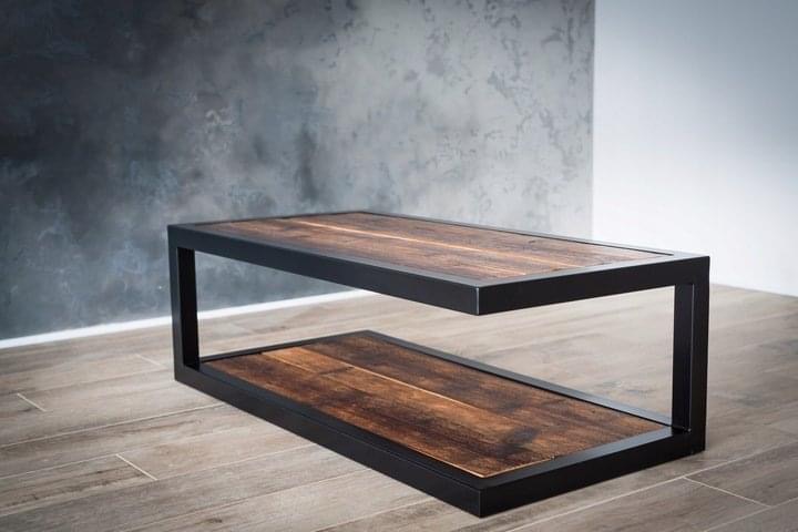 Stolik kawowy w stylu industrialnym z ciemnego drewna i czarnej, metalowej ramy, dwupoziomowy, na tle szarej ściany imitującej beton.