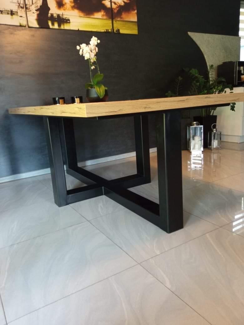 Stół z drewnianym blatem i czarną, metalową podstawą w kształcie litery X, wykonany w Nieczajnej Górnej. Na blacie stoi doniczka z białą orchideą.