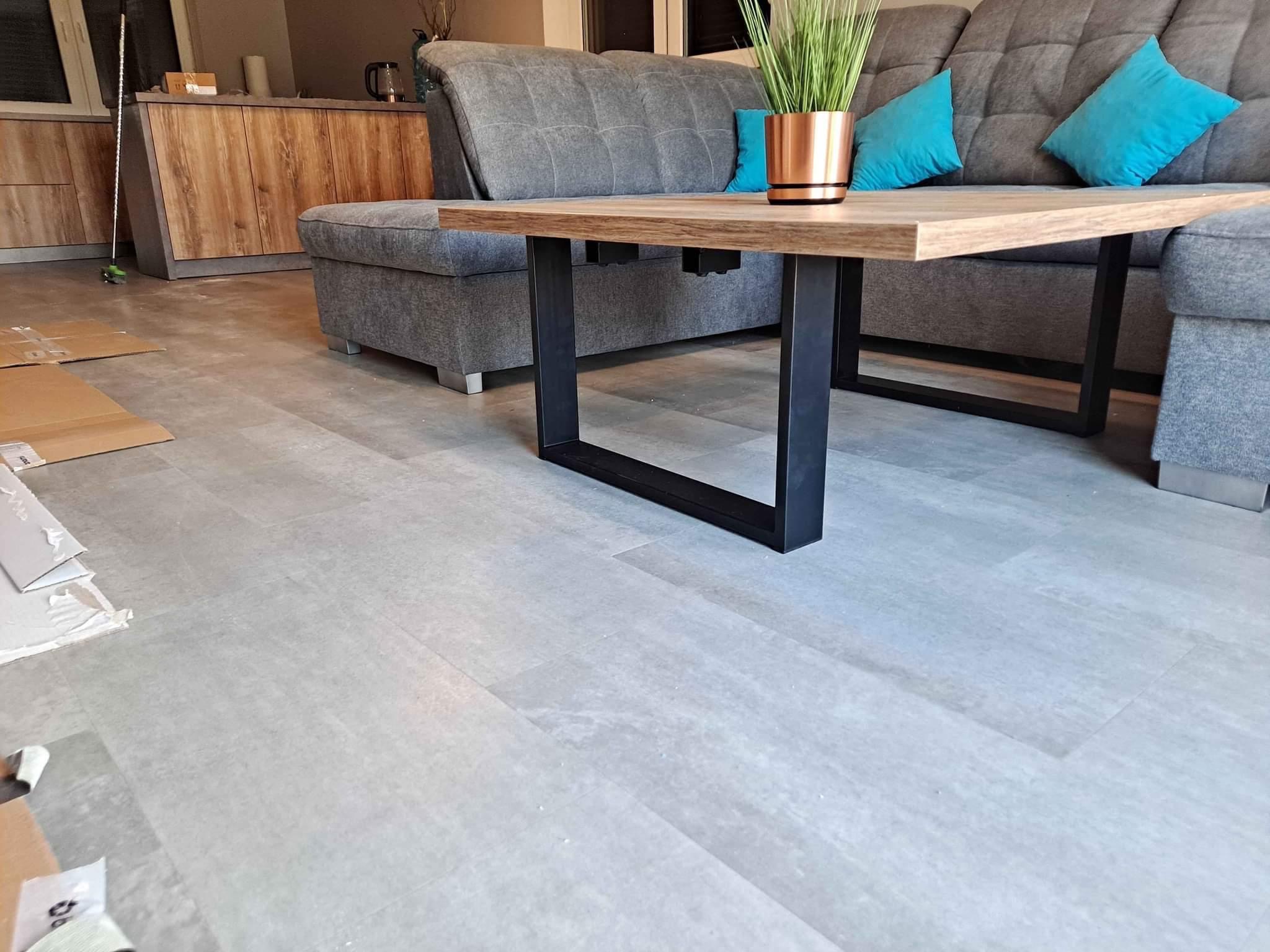 Nowoczesny stolik kawowy z drewnianym blatem i czarnymi, prostokątnymi nogami, ustawiony na jasnoszarej podłodze w salonie z szarą sofą i turkusowymi poduszkami w tle. Na blacie stoi doniczka...
